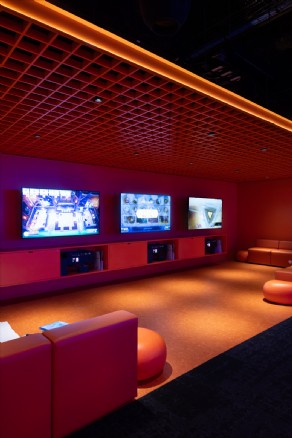 Lounge area illuminated by ambient gaming lights with orange ceiling grid and screen displays at Syracuse University.